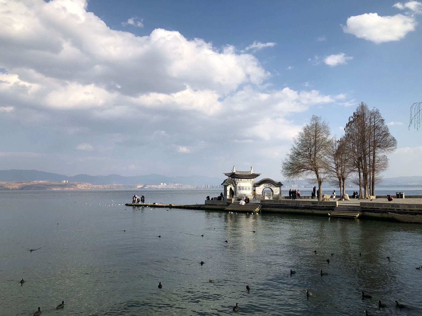 Dianchi Lake at Kunming: a stone promenade with a moon-gate pavilion and bare trees along the shore, waterbirds on the calm lake, distant hills and a pagoda under a big cloudy sky