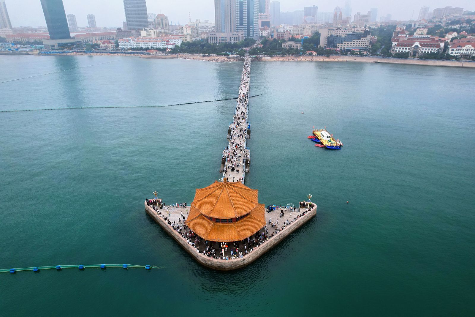 Aerial view of Zhanqiao Pier stretching into the sea at Qingdao, ending at the orange-tiled octagonal Huilan Pavilion, with the red-roofed old town skyline behind